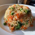 A beautifully molded serving of Vegetable Fried Rice with Sesame Ginger Sauce, featuring carrots, peas, and broccoli, topped with sesame seeds.
