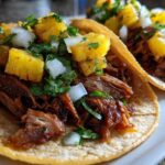 Close-up of two Al Pastor Pork Tacos topped with chunks of pineapple, diced white onion, and cilantro.