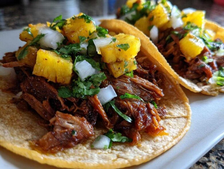 Close-up of two Al Pastor Pork Tacos topped with chunks of pineapple, diced white onion, and cilantro.