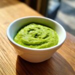 A close-up of bright green Avocado Tomatillo Salsa Verde swirled in a small white bowl on a wooden surface.