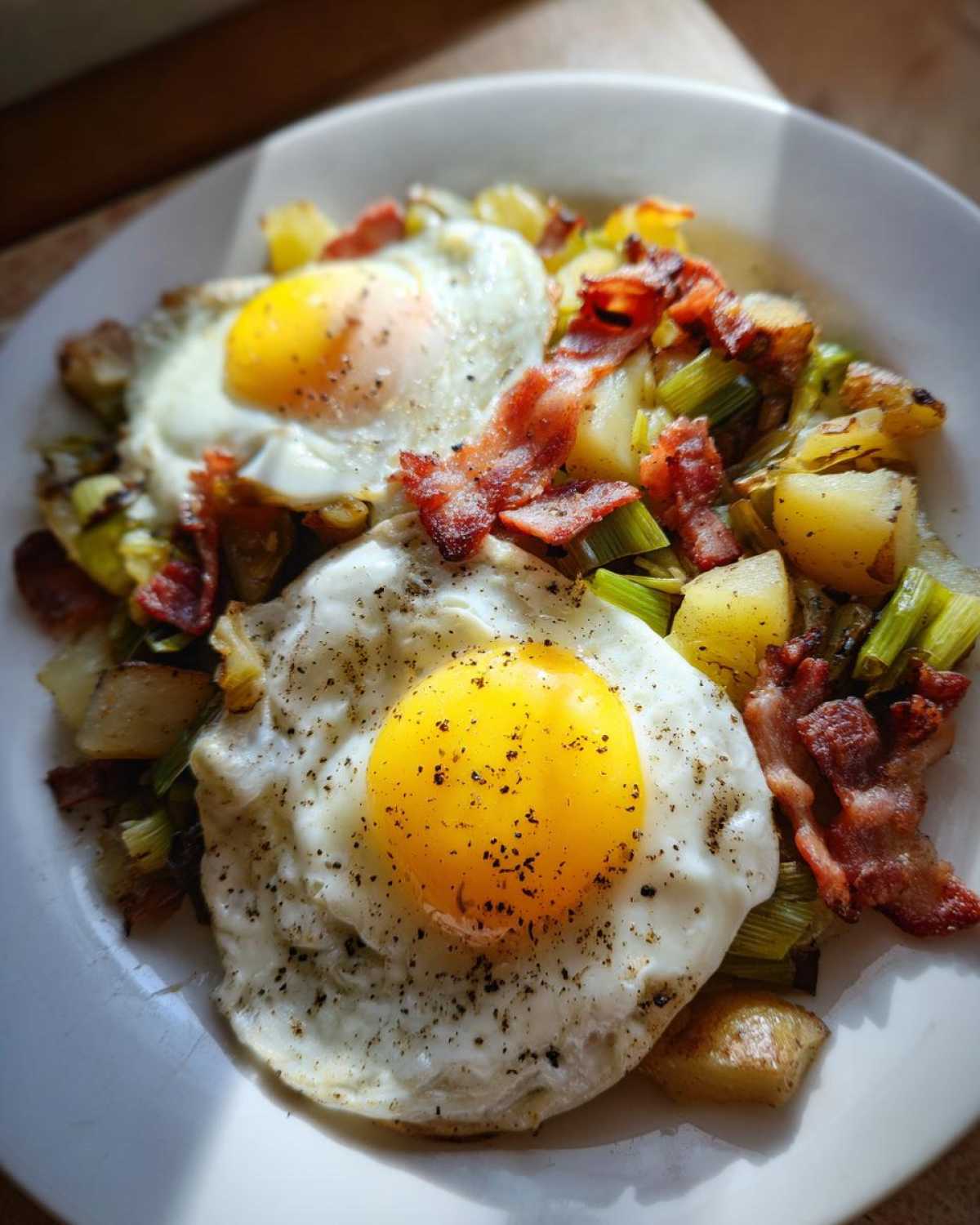 A plate of Bacon Leek Breakfast Hash topped with two perfectly cooked sunny-side-up eggs and cracked pepper.