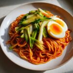 A plate of vibrant Bibim Guksu Spicy Cold Noodles tossed in red sauce, topped with julienned cucumbers and a soft-boiled egg.