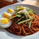 A close-up of a bowl of Bibim Guksu Spicy Cold Noodles topped with julienned cucumber and sliced hard-boiled eggs.