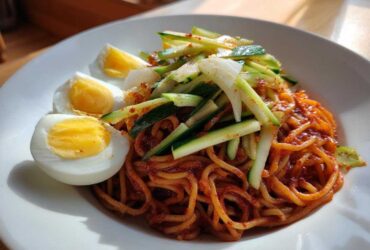 A close-up of a bowl of Bibim Guksu Spicy Cold Noodles topped with julienned cucumber and sliced hard-boiled eggs.