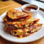 A stack of three crispy, cheesy Birria Quesatacos served on a white plate next to a small cup of dipping consome.