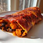 A single serving of Black Bean Sweet Potato Enchiladas covered in red sauce, showing the orange sweet potato and black bean filling.