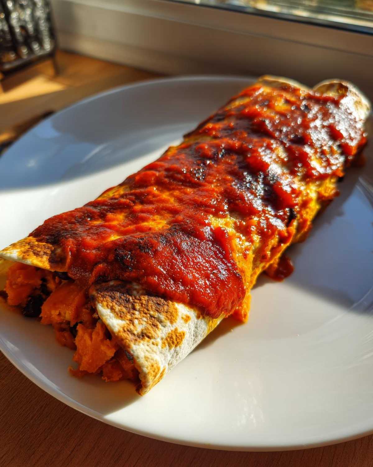 A single, rolled Black Bean Sweet Potato Enchiladas covered in vibrant red sauce, served on a white plate.