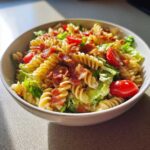 A close-up of a white bowl filled with BLT Pasta Salad with Ranch, featuring rotini pasta, lettuce, tomatoes, and crispy bacon.