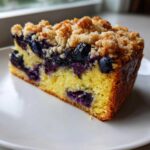 A close-up of a moist slice of Blueberry Almond Coffee Cake featuring a yellow crumb, bursting blueberries, and a crunchy streusel topping.