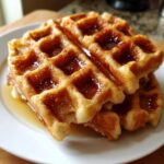 Close-up of stacked, golden Brown Sugar Banana Waffles drizzled generously with syrup on a white plate.