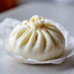 A single, perfectly steamed Char Siu Mushroom Bao bun topped with sesame seeds, resting on white parchment paper.