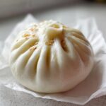 Close-up of a single, fluffy, white Char Siu Mushroom Bao sprinkled with toasted sesame seeds.
