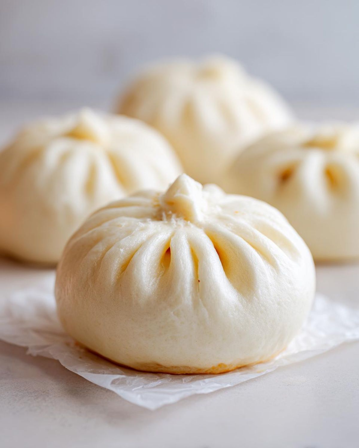A close-up of a fluffy, white steamed Char Siu Mushroom Bao resting on parchment paper, with more buns blurred in the background.
