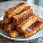 A close-up stack of golden brown Churro French Toast Sticks heavily coated in sparkling cinnamon sugar, served on a white plate.