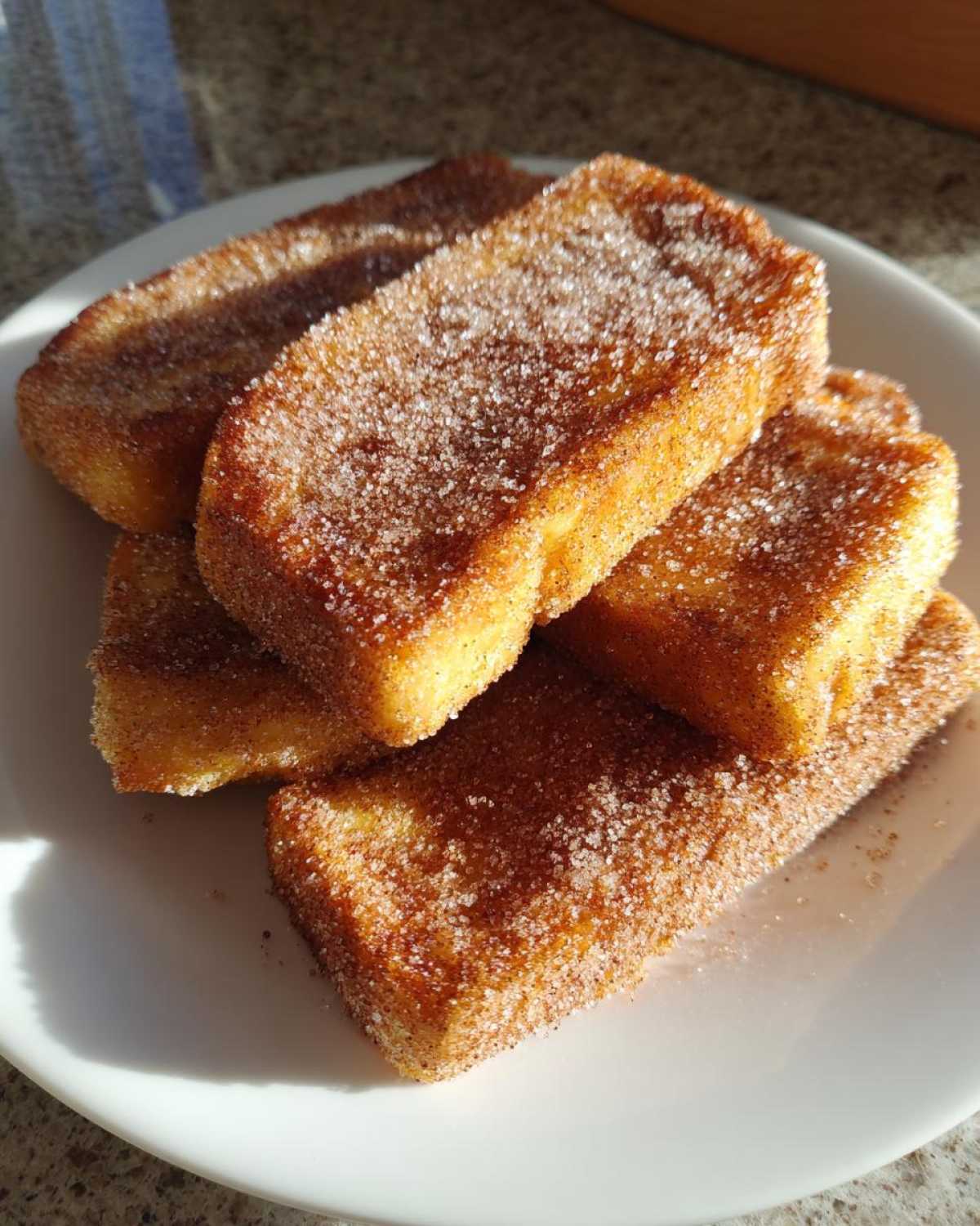 A stack of golden brown Churro French Toast Sticks heavily coated in cinnamon sugar, resting on a white plate.