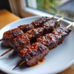 Close-up of grilled Coffee Rub Flank Steak Skewers glazed with a spicy sauce on a white plate.