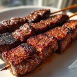 Close-up of perfectly grilled Coffee Rub Flank Steak Skewers resting on a white plate, showing a rich, dark crust.