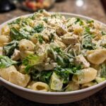 A close-up of a white bowl filled with Cold Chicken Caesar Pasta Salad featuring shell pasta, chicken chunks, romaine lettuce, and creamy dressing.
