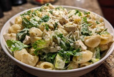 A close-up of a white bowl filled with Cold Chicken Caesar Pasta Salad featuring shell pasta, chicken chunks, romaine lettuce, and creamy dressing.
