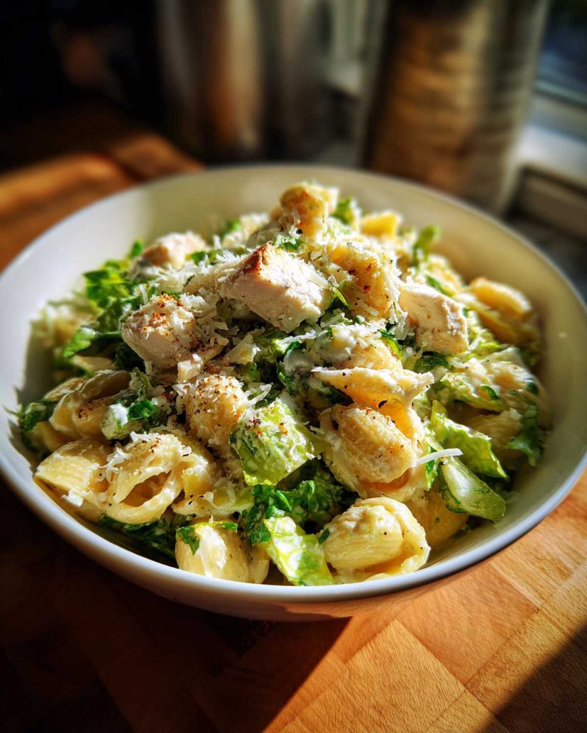 A close-up of Cold Chicken Caesar Pasta Salad in a white bowl, featuring pasta shells, chicken pieces, romaine lettuce, and grated Parmesan.