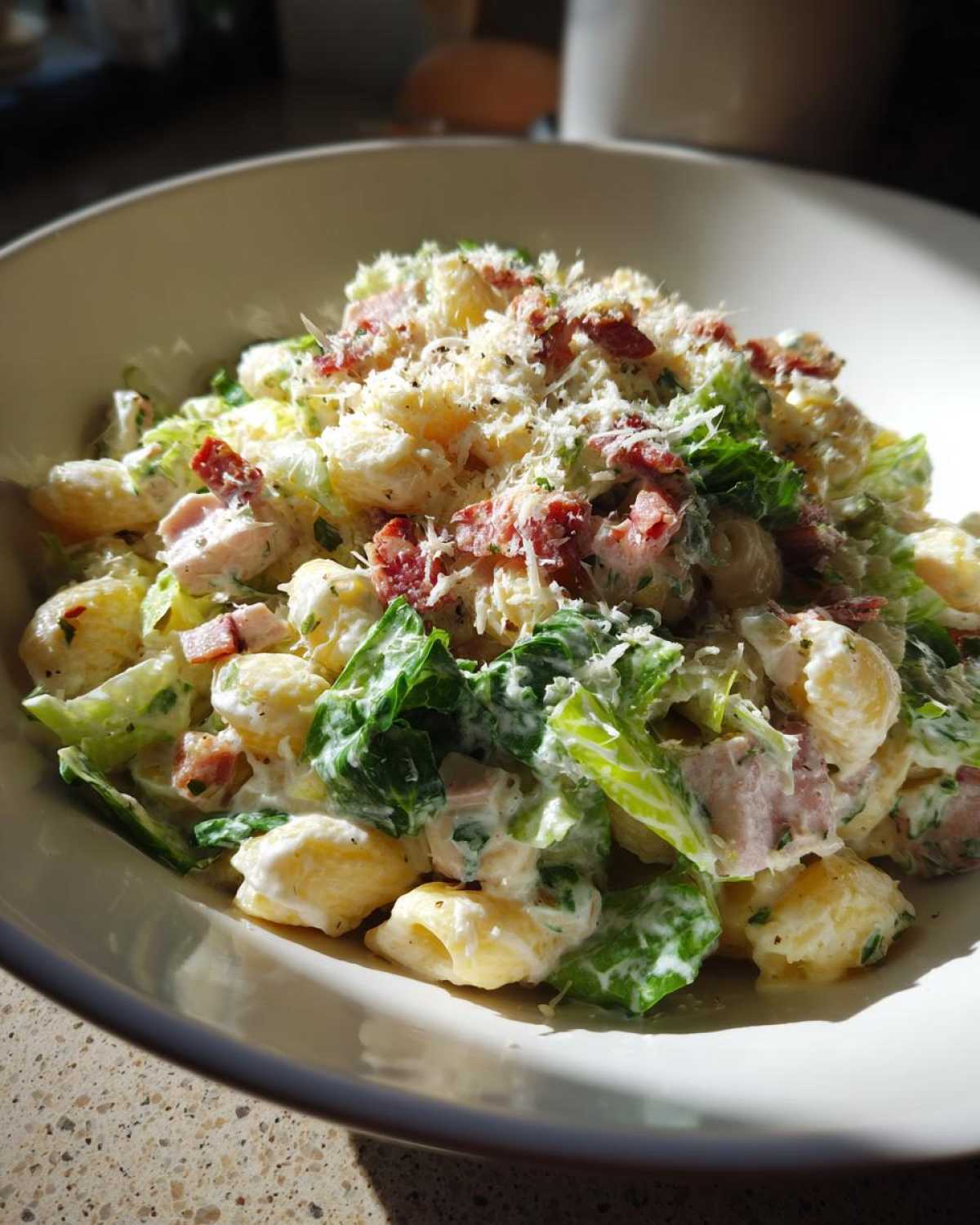 A close-up of a bowl filled with Cold Chicken Caesar Pasta Salad, featuring pasta, creamy dressing, romaine lettuce, bacon bits, and grated cheese.