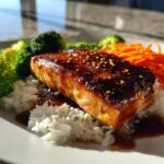 A perfectly cooked fillet of salmon with a dark, glossy glaze and sesame seeds, served over white rice with steamed broccoli and shredded carrots for Crispy Salmon Rice Bowls.