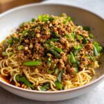 A close-up of a bowl of Dan Dan Noodles with Sesame, featuring noodles tossed in chili oil and topped with savory ground meat, green onions, and crushed peanuts.