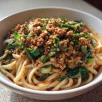 Close-up of a white bowl filled with Dan Dan Noodles with Sesame, topped with ground meat, greens, and chopped peanuts.