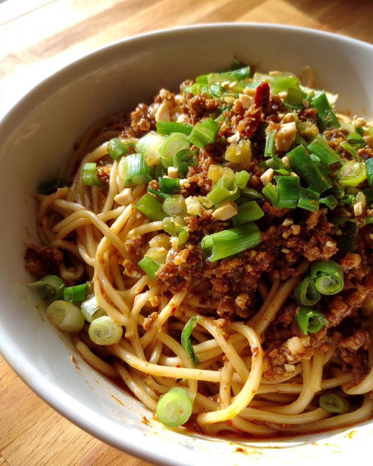 A close-up of rich Dan Dan Noodles with Sesame topped with savory ground meat, chopped scallions, and crushed peanuts.