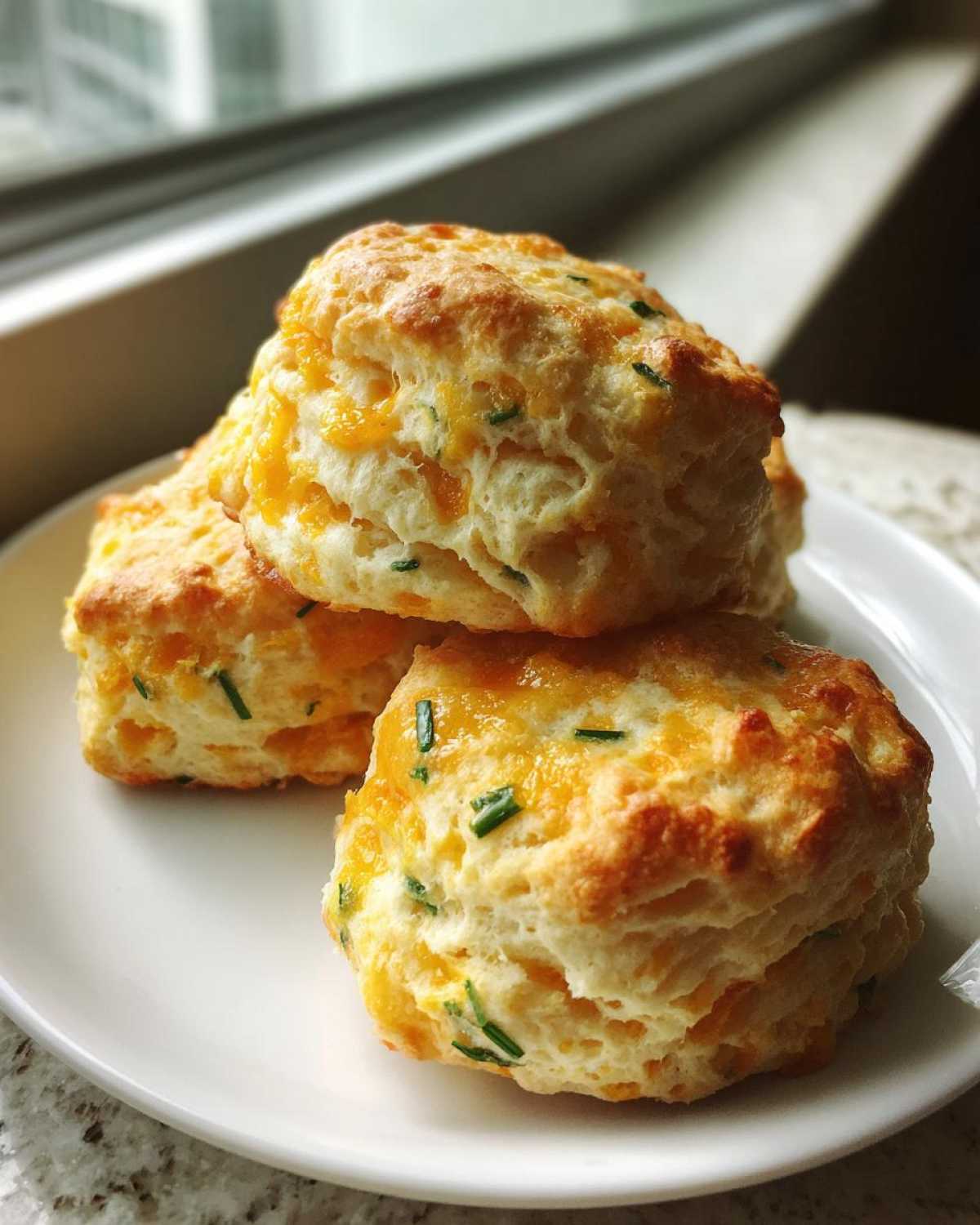 Three golden brown, fluffy Chive Cheddar Biscuits stacked on a white plate near a window.