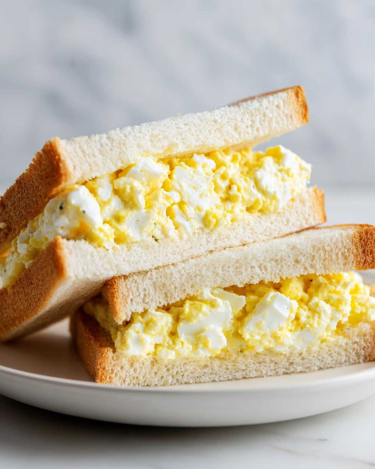 Two halves of a Tamago Sando Japanese Egg Sandwich stacked on a white plate, showing creamy egg salad filling.