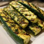 Plump slices of Grilled Zucchini with Lemon Herb Marinade showing distinct char marks, piled on a white serving dish.