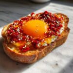 A close-up of Jammy Egg Toast with Chili Crisp, featuring a bright orange yolk and red chili flakes on toasted bread.