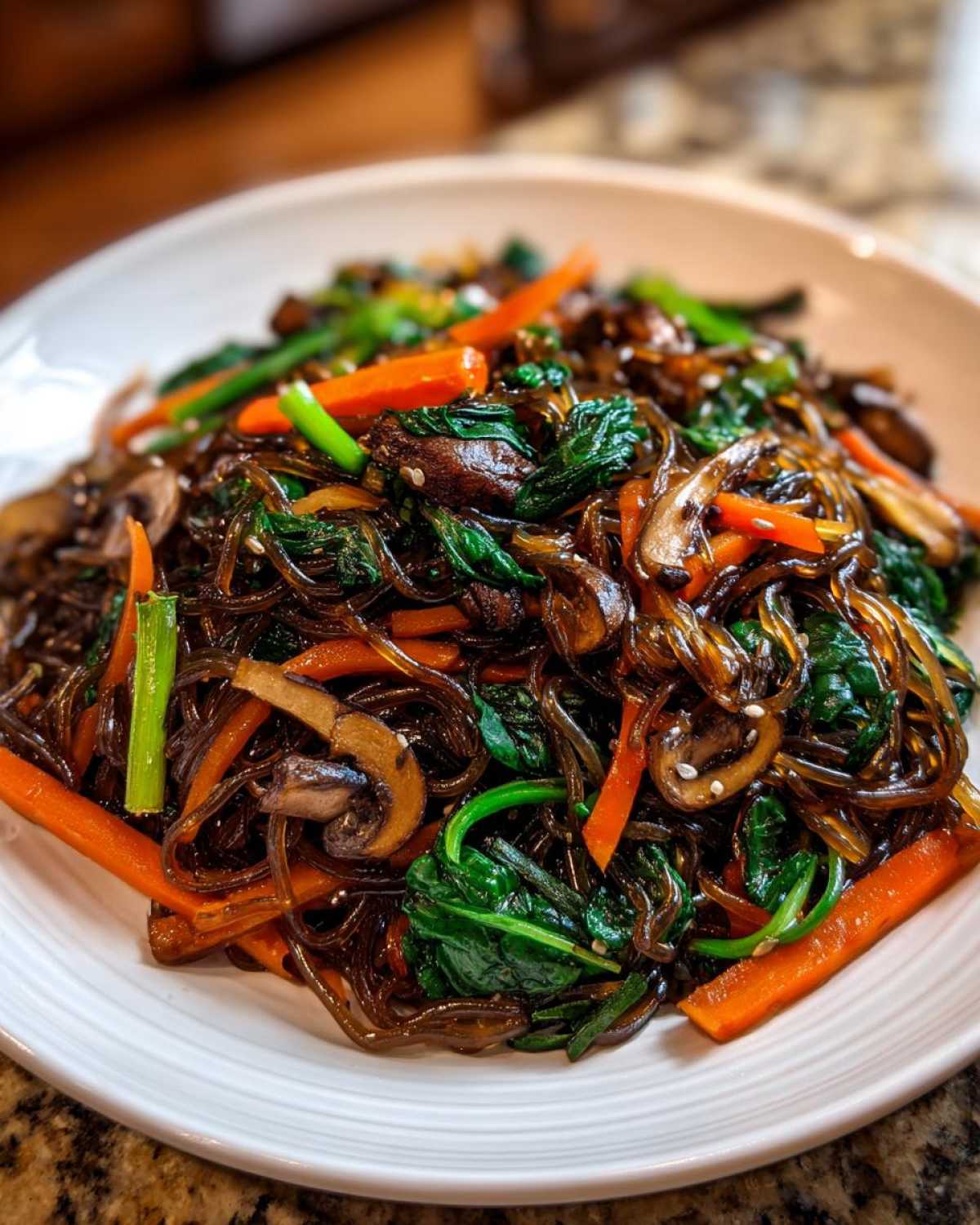 A close-up serving of glossy Japchae Sweet Potato Noodles mixed with vibrant spinach, sliced carrots, and mushrooms.