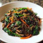 A close-up of savory Japchae sweet potato noodles mixed with dark green spinach, bright orange carrots, and sliced shiitake mushrooms.