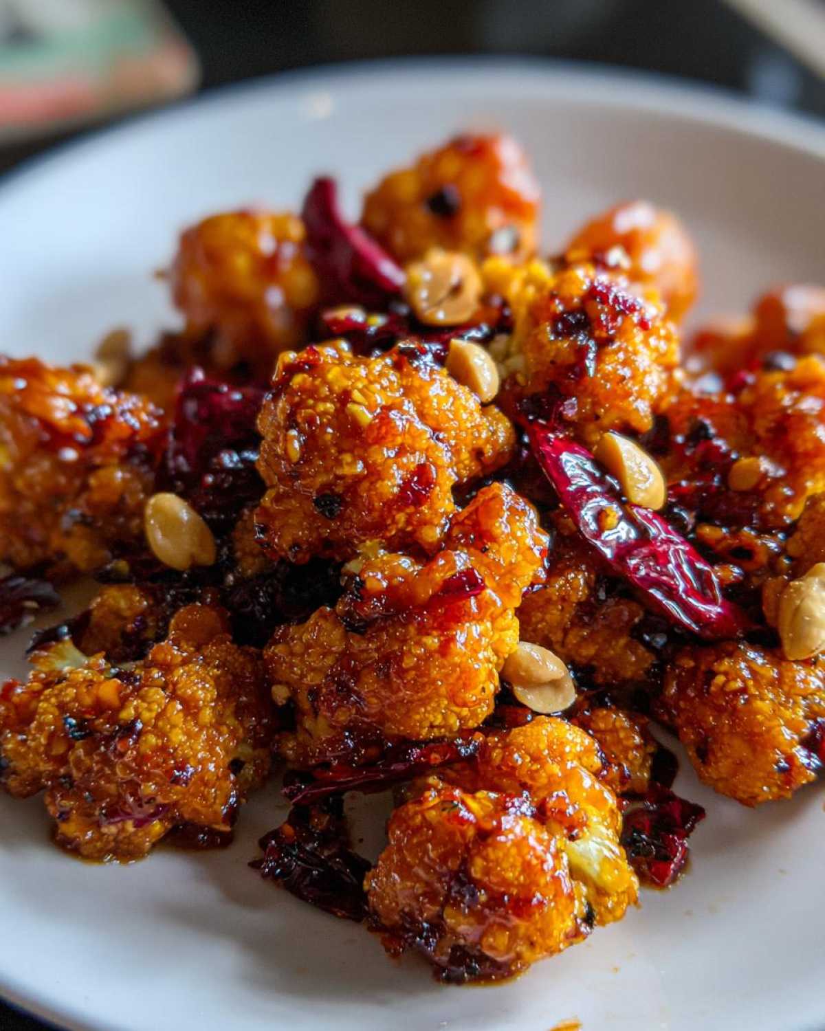 Close-up of crispy, glazed Kung Pao Cauliflower florets mixed with whole dried chilies and peanuts on a white plate.
