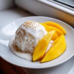 A serving of Mango Sticky Rice with Coconut, featuring sticky rice drizzled with coconut sauce and topped with sesame seeds, next to bright yellow mango slices.