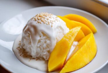 A serving of Mango Sticky Rice with Coconut, featuring sticky rice drizzled with coconut sauce and topped with sesame seeds, next to bright yellow mango slices.