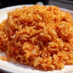 Close-up of perfectly cooked Mexican Rice with Roasted Garlic, showing individual orange-red grains piled on a white plate.