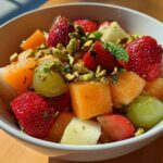 Close-up of a white bowl filled with Orange Blossom Fruit Salad featuring strawberries, cantaloupe, grapes, and chopped pistachios.