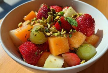 Close-up of a white bowl filled with Orange Blossom Fruit Salad featuring strawberries, cantaloupe, grapes, and chopped pistachios.