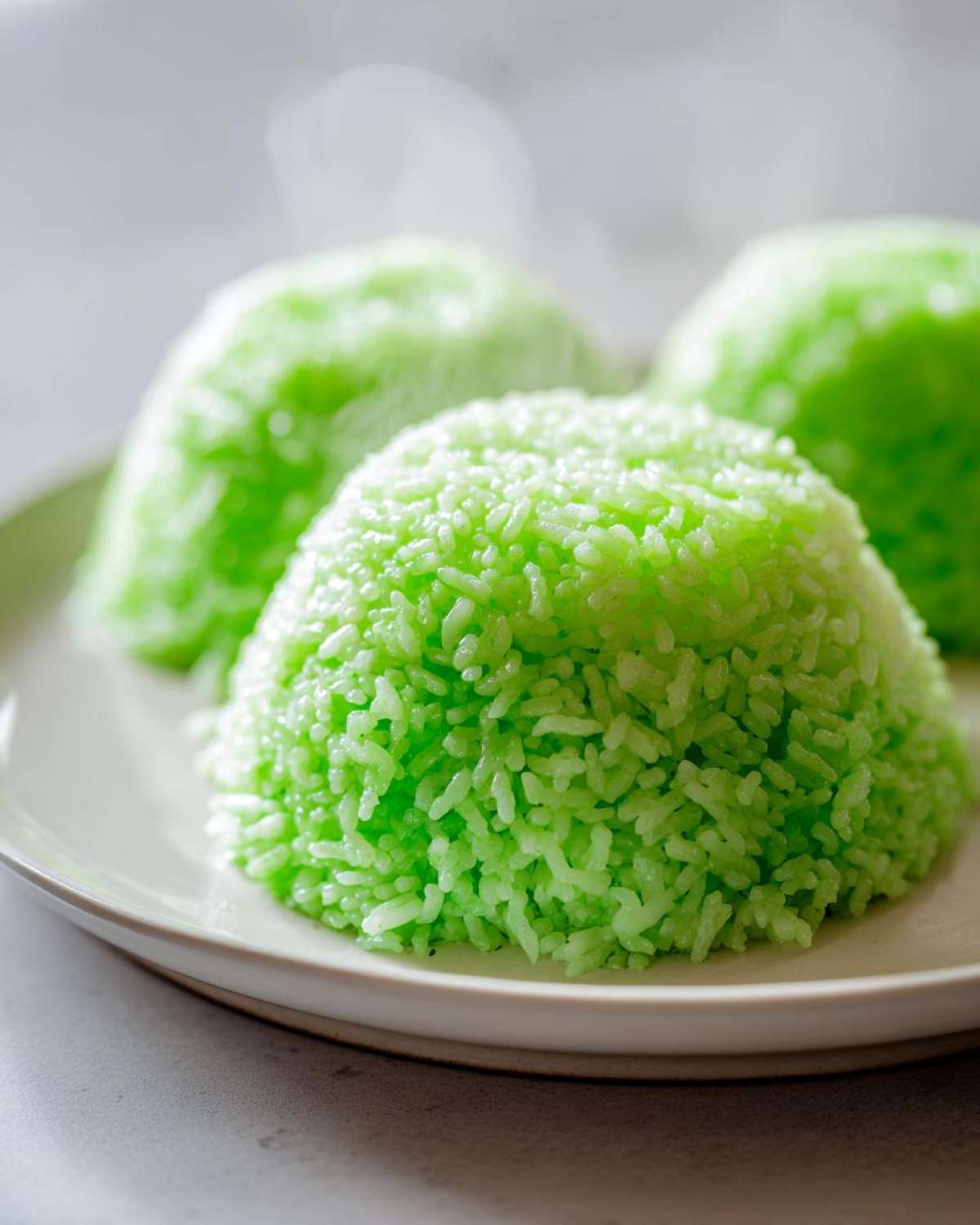 Three molded servings of vibrant green Pandan Coconut Rice steaming slightly on a light ceramic plate.