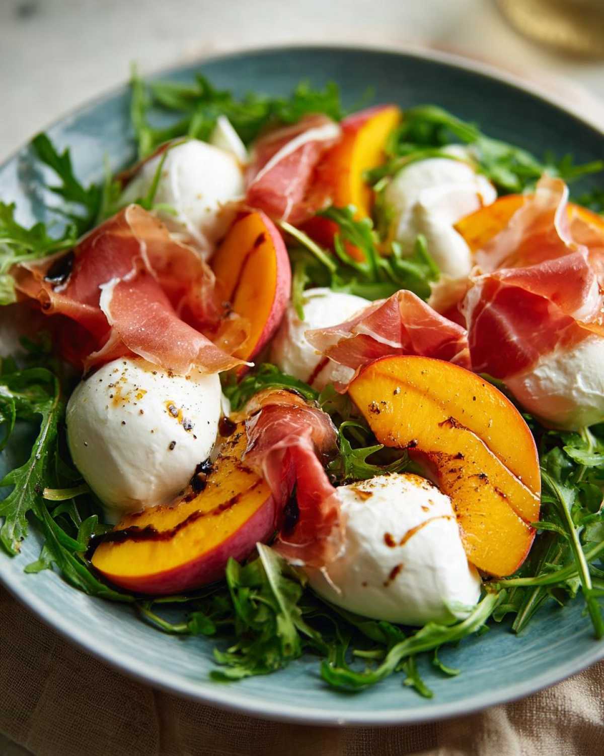 Close-up of a Peach Burrata Prosciutto Salad featuring grilled peaches, fresh burrata, and prosciutto over arugula, drizzled with balsamic glaze.