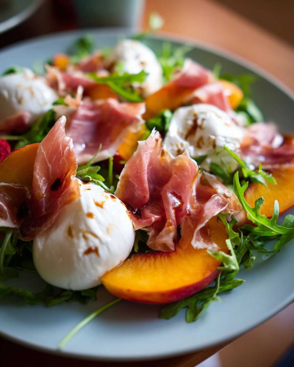 Close-up of a vibrant Peach Burrata Prosciutto Salad featuring fresh burrata balls, sliced peaches, prosciutto, and arugula drizzled with balsamic.