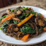 A close-up of a serving of Japchae Sweet Potato Noodles mixed with spinach, carrots, and mushrooms on a white plate.