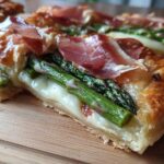 A cross-section view of a slice of Prosciutto Asparagus Tart showing flaky pastry, melted cheese, bright green asparagus, and draped prosciutto.