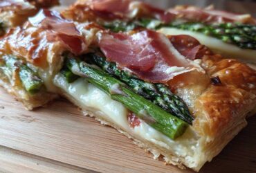 A cross-section view of a slice of Prosciutto Asparagus Tart showing flaky pastry, melted cheese, bright green asparagus, and draped prosciutto.