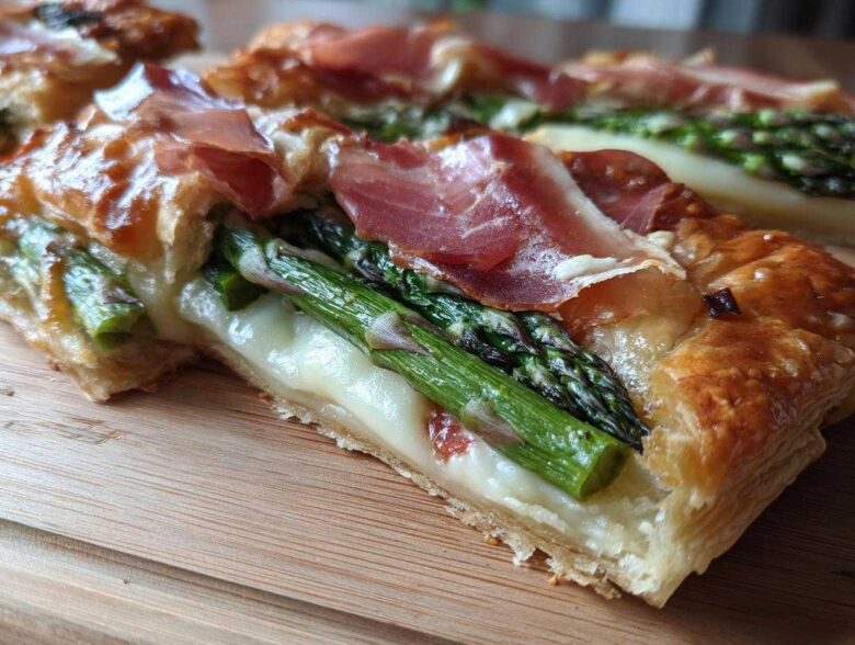 A cross-section view of a slice of Prosciutto Asparagus Tart showing flaky pastry, melted cheese, bright green asparagus, and draped prosciutto.