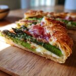 Close-up of a slice of Prosciutto Asparagus Tart showing layers of green asparagus, melted cheese, and prosciutto on a golden, flaky pastry crust.