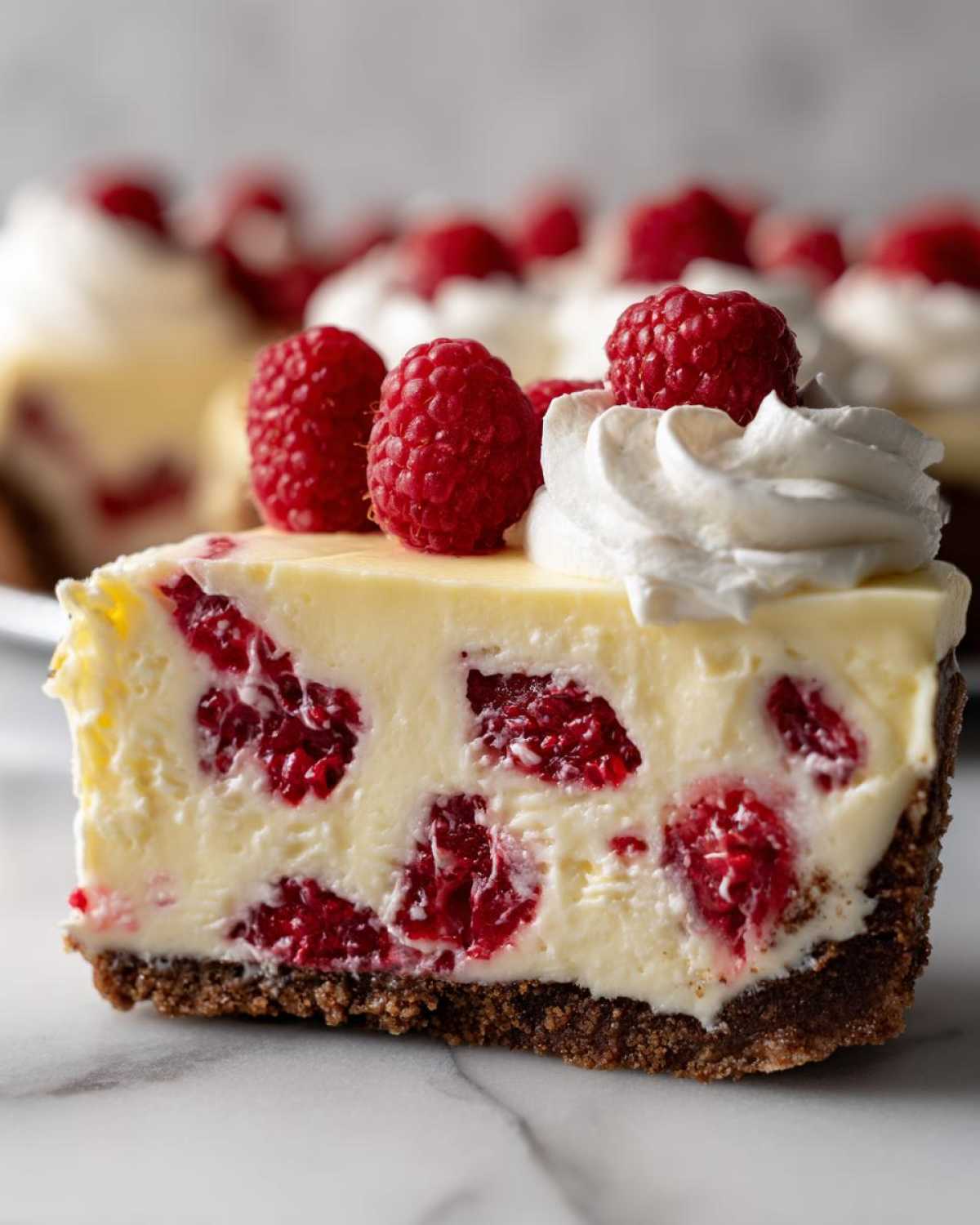 Close-up of a slice of Raspberry Lemonade Icebox Pie showing a creamy filling with whole raspberries, a dark crust, and whipped cream topping.